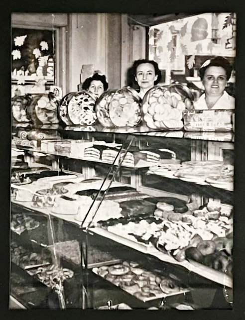 Three white women standing behind glass display case filled with baked goods