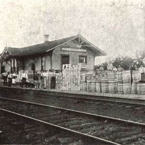 People and various barrels and other items around single story wooden building beside railroad tracks