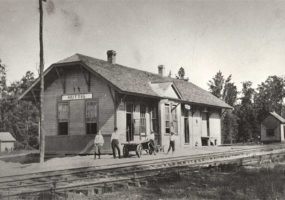 Four men standing before single story wooden building beside railroad tracks