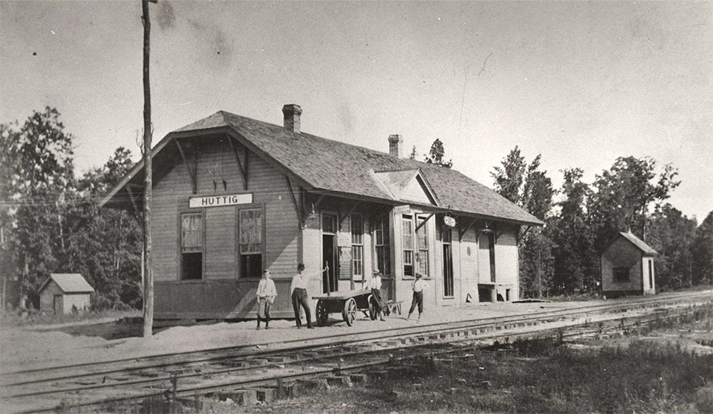 Four men standing before single story wooden building beside railroad tracks