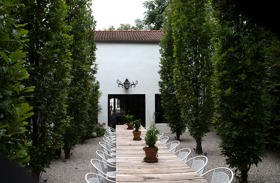 Patio with long table and chairs on each side amid trees and white building
