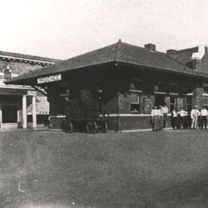 Group of men standing before single story building next to railroad tracks