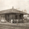 Group of men in front of single story wooden building beside railroad tracks