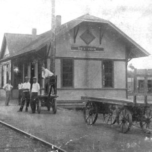 Single story building next to railroad tracks with people milling about and man sitting by tracks with dog