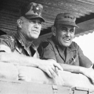 Three white men in military garb leaning on wall beneath sheet metal roof