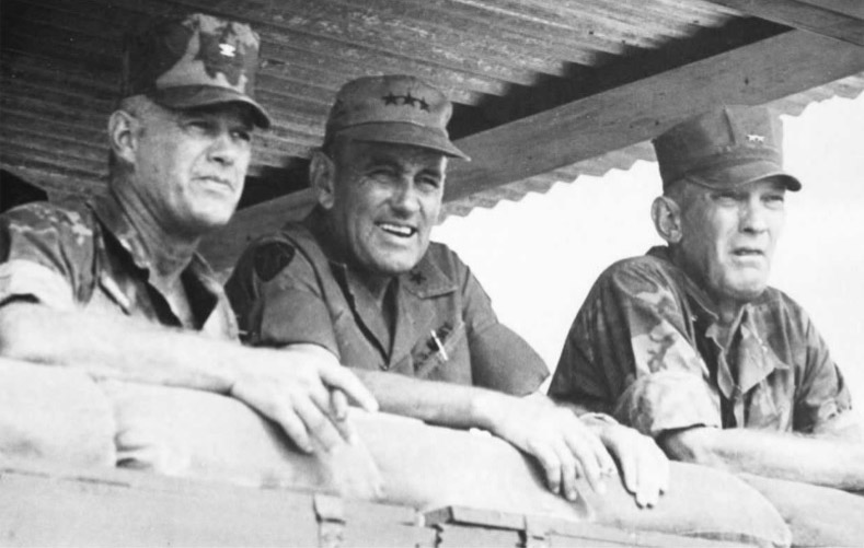 Three white men in military garb leaning on wall beneath sheet metal roof