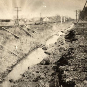 Railroad tracks running beside ditch; houses in the background