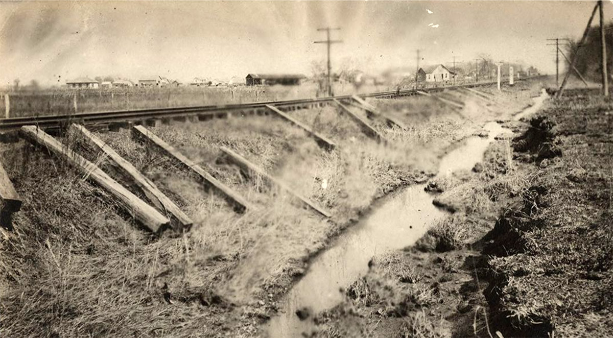 Railroad tracks running beside ditch; houses in the background