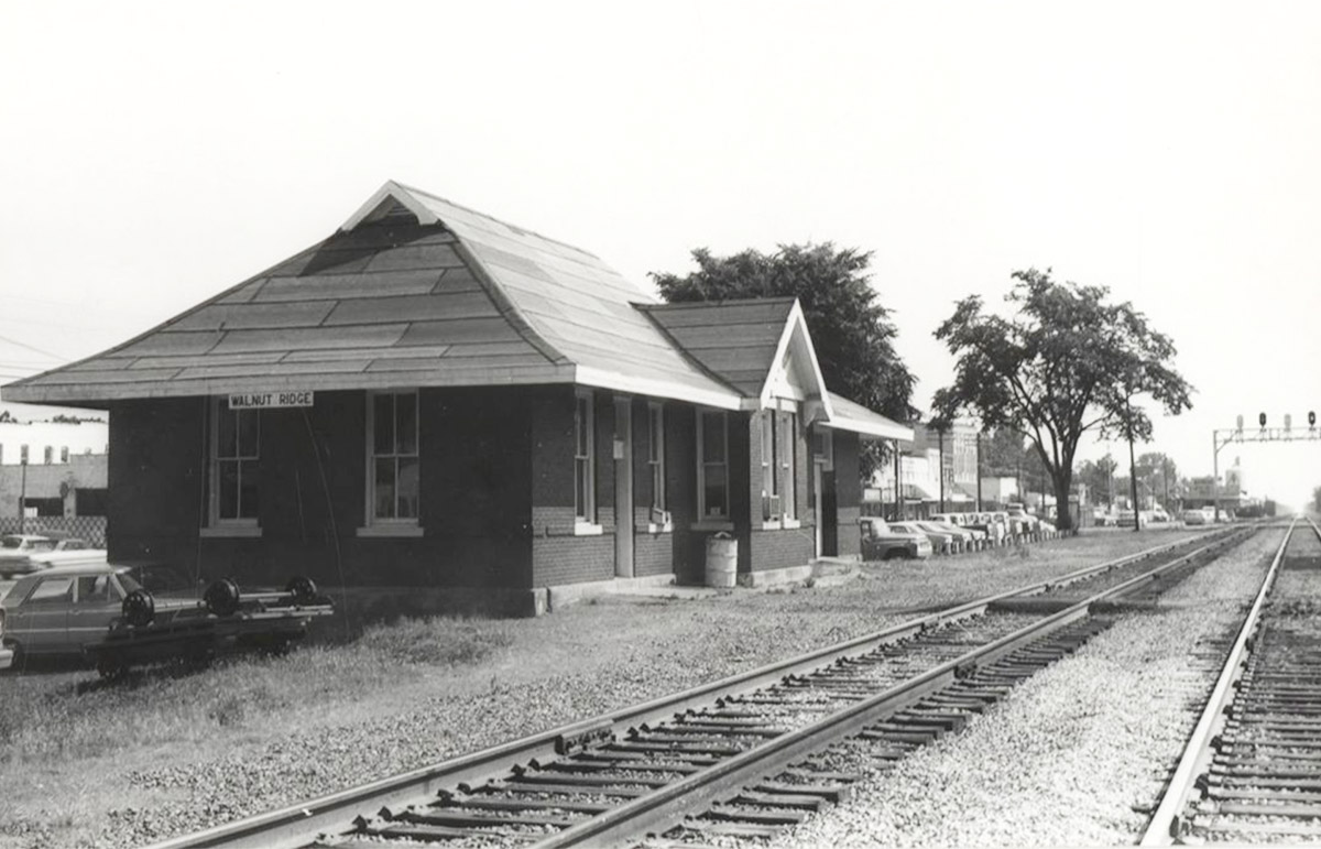 Single story brick building beside railroad tracks with a line of cars parked next to it