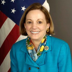 White woman in blue suit and scarf in front of American flag