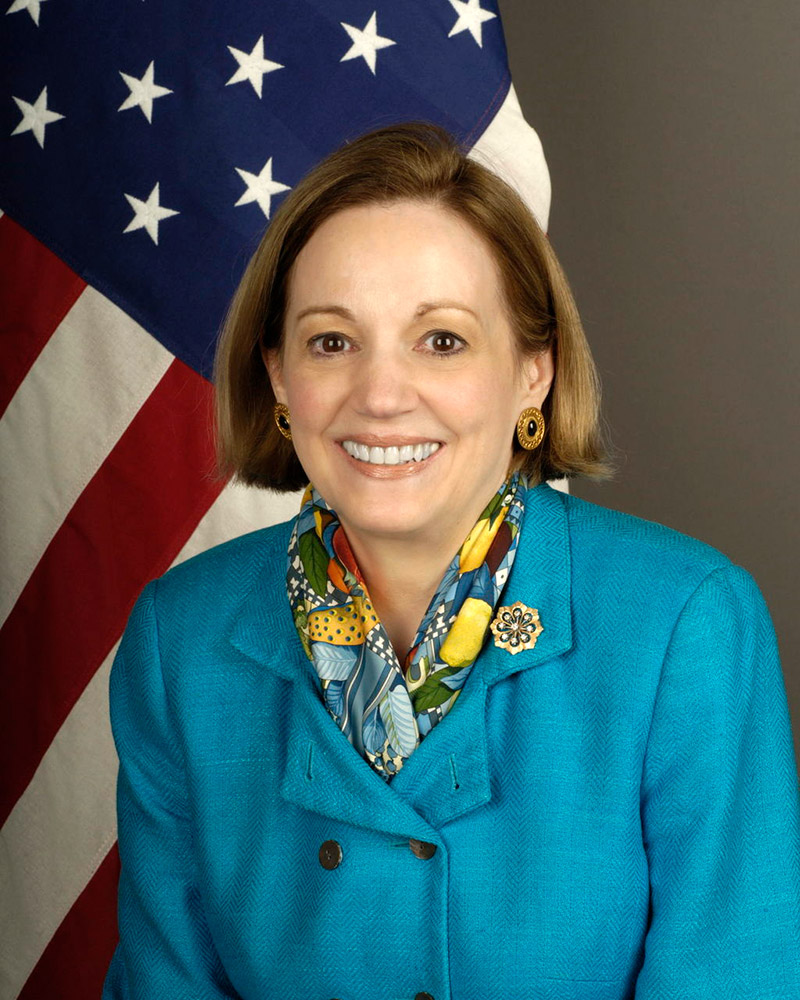 White woman in blue suit and scarf in front of American flag