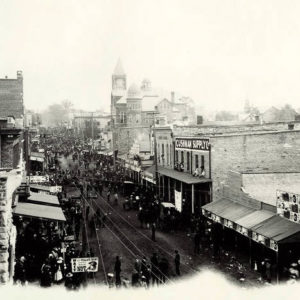 City street seen from elevated position with multistory storefronts along road lined with wires and filled with people