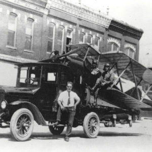 One man standing beside truck and two men in the cockpit of the plane on the back of the truck