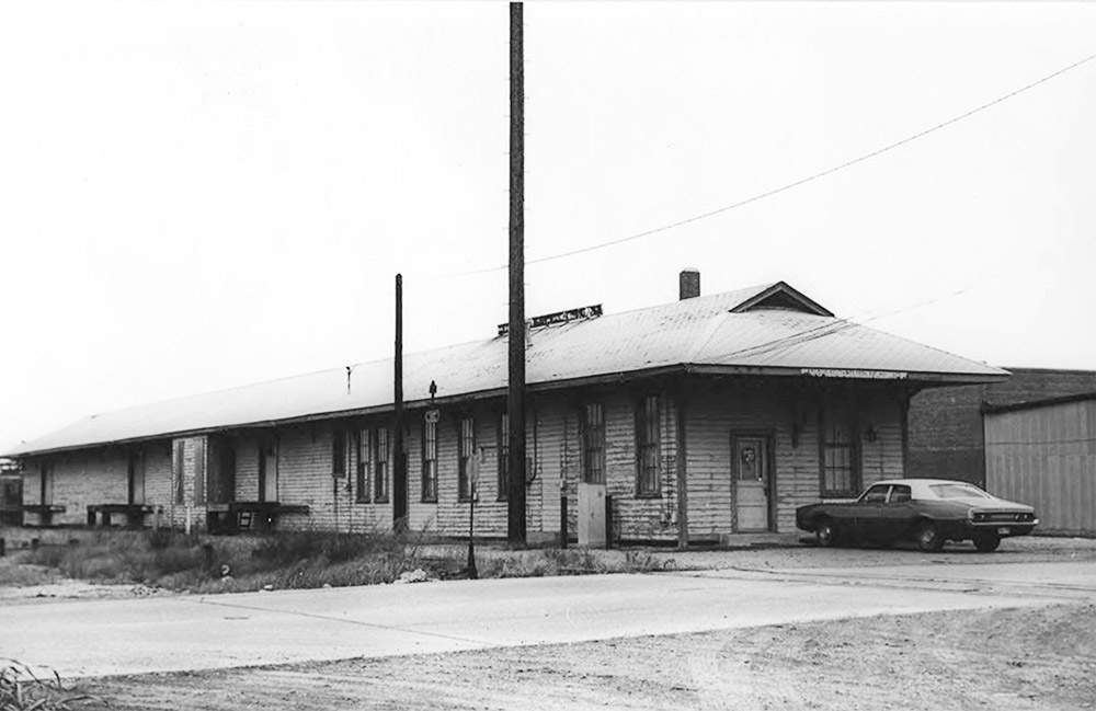 Single story wooden building next to railroad tracks with car parked at the end