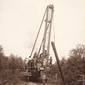 Men working a derrick lifting a pole on a railroad car