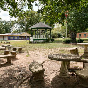 Gazebo and concrete picnic tables on patchy grass with buildings in the background