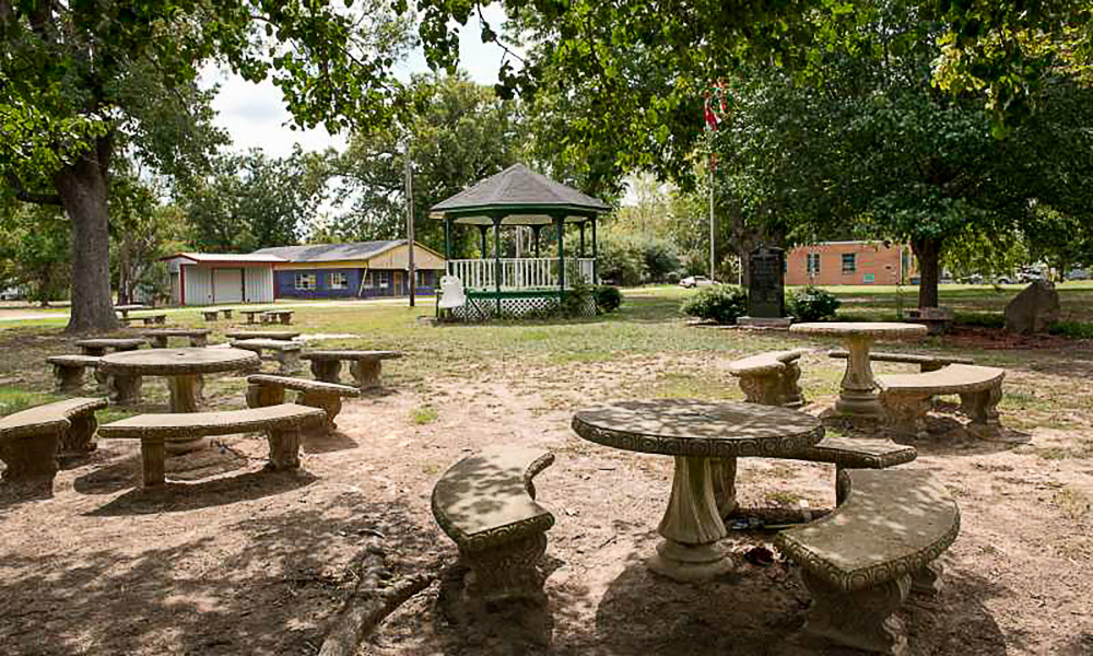 Gazebo and concrete picnic tables on patchy grass with buildings in the background