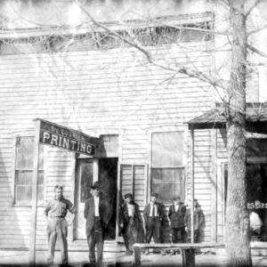 Six white people standing in front of multistory wooden building with sign saying "Courier Printing"
