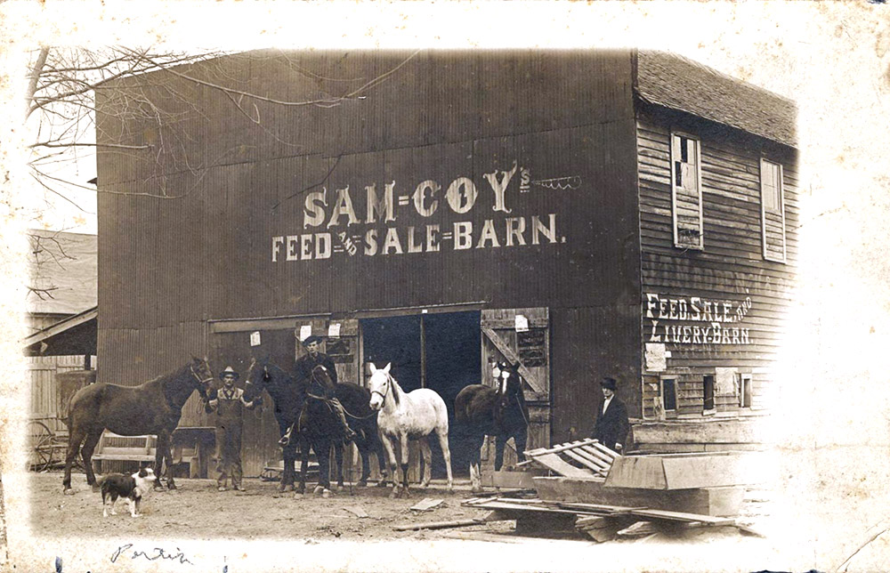 Multiple story wooden buildings with man and horses standing in front "Sam Coy Feel and Sale Barn"