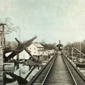 Train on tracks leading to bridge with dredge boats on either side of bridge