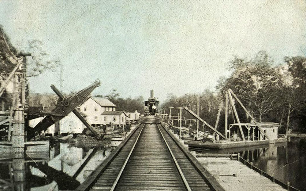 Train on tracks leading to bridge with dredge boats on either side of bridge