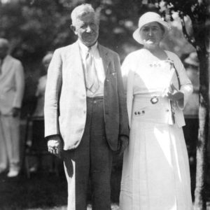White man in suit and woman in white dress and hat standing outdoors
