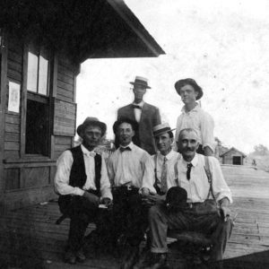 six white men seated and standing on wooden porch adjacent to wooden building