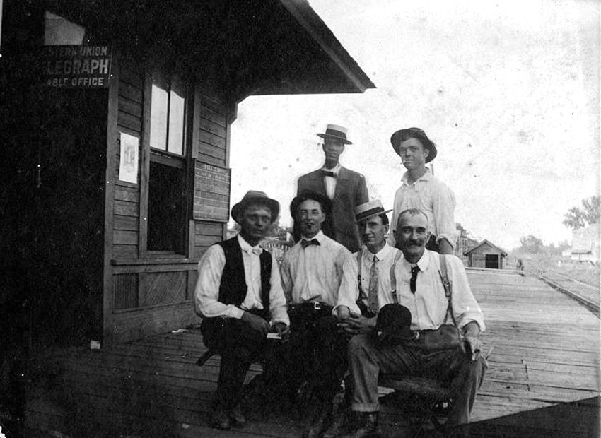 six white men seated and standing on wooden porch adjacent to wooden building