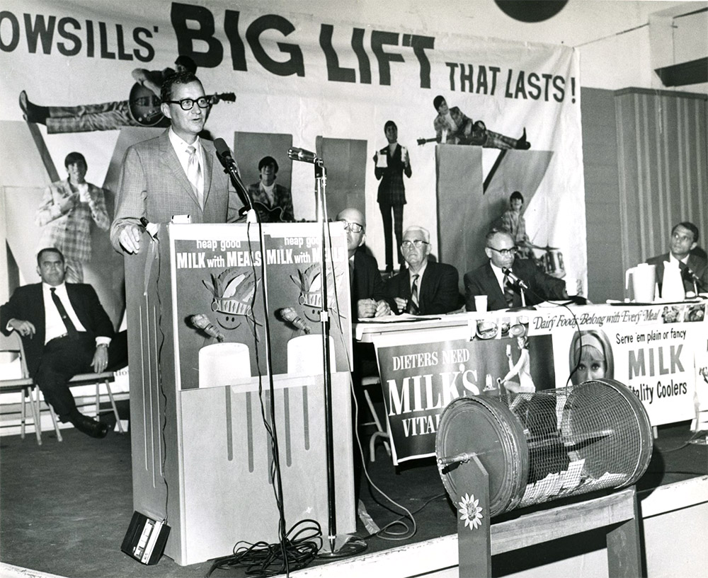 White man in suit speaking at lectern in front of promotional signs