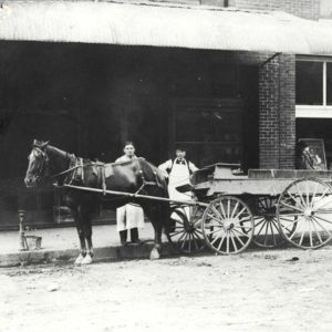 White men in aprons standing horse and wagon in front of brick multistory building.