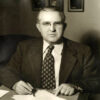 White man in glasses and suit holding pen above papers on table