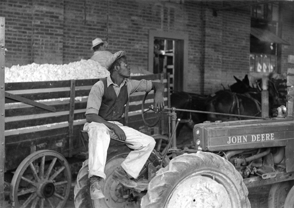 African American man sitting on tractor beside wagon loaded with cotton and brick building in background