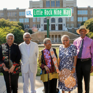 Five African Americans standing beneath a sign "Little Rock Nine Way" in front of a large brick school building