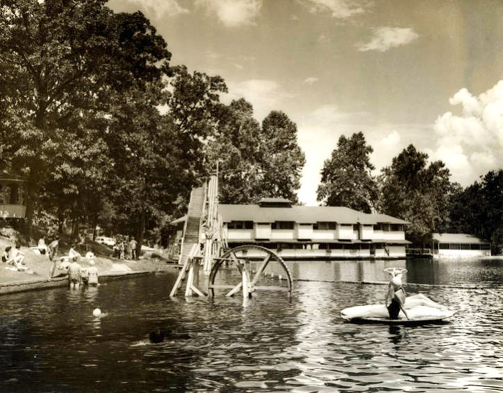Tree-lined lake short with multistory building in distance and people playing in the water