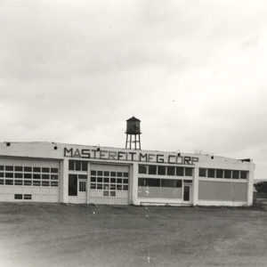 Single story white concrete block building with water tank on top with "Masterfit Manufacturing Corporation" on the front