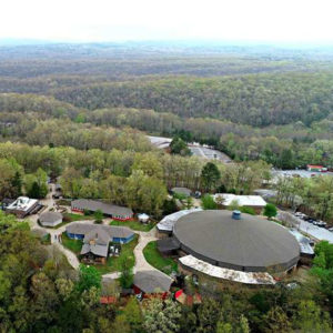 Overhead view of large round building and adjacent buildings and trees and roads and cars