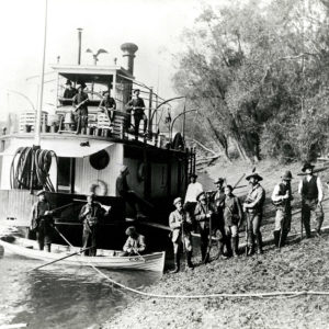 Large group of men with guns and dogs beside steamship moored at riverbank