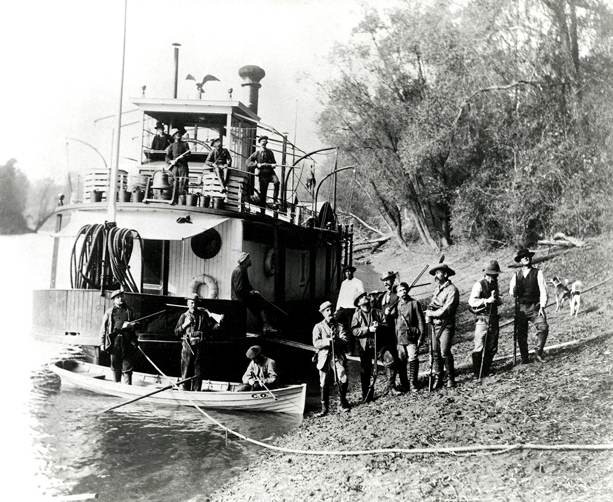 Large group of men with guns and dogs beside steamship moored at riverbank
