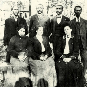 Three African American women in blouses and skirts seated; four suited African American men standing behind them