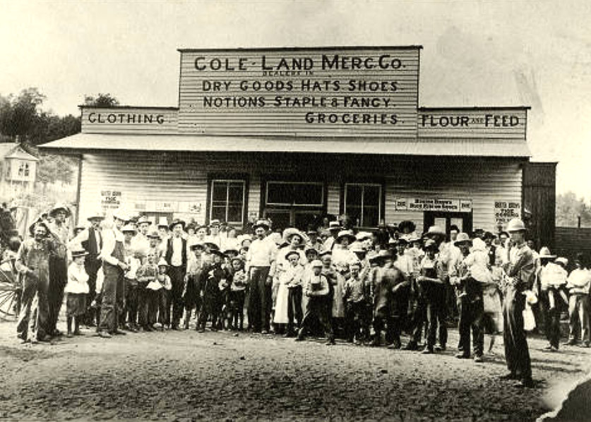 Large group of people standing in front of wooden building "Cole Land Mercantile"