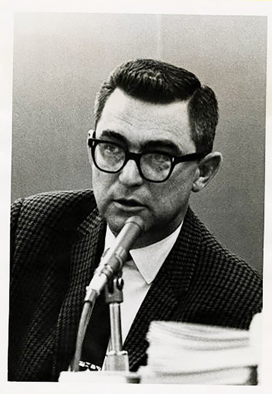 White man in suit and black-framed glasses speaking into microphone
