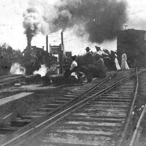 Group of people sitting and standing beside railroad tracks with steam locomotive approaching