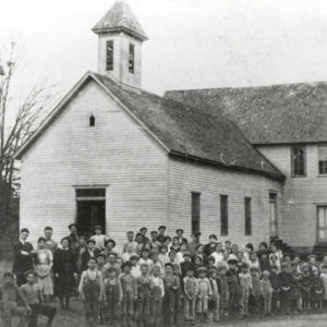 Large group of children and adults gathered in front of white multistory wooden building