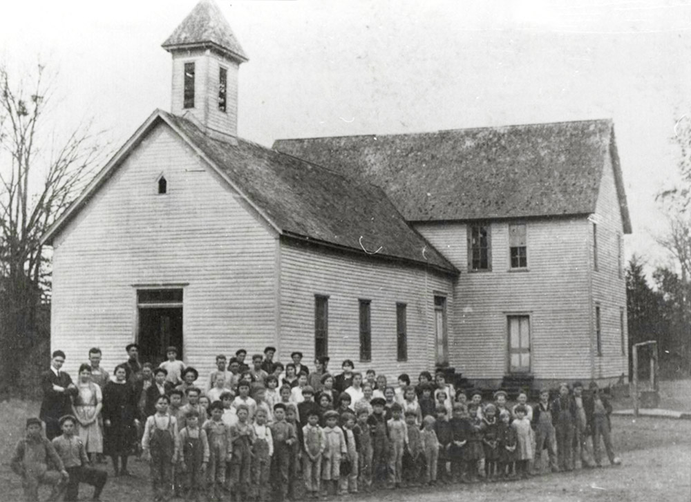 Large group of children and adults gathered in front of white multistory wooden building