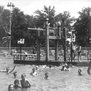 Many white children swimming in large pool with diving board and lifeguard seat