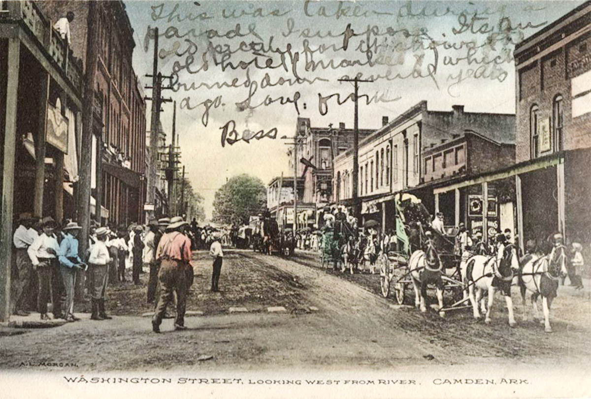 Large crowd of people on street with horses and wagons and multistory storefront buildings on both sides