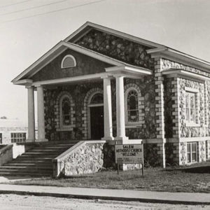 Single story rock church building with steps leading up to entrance a car parked next to it