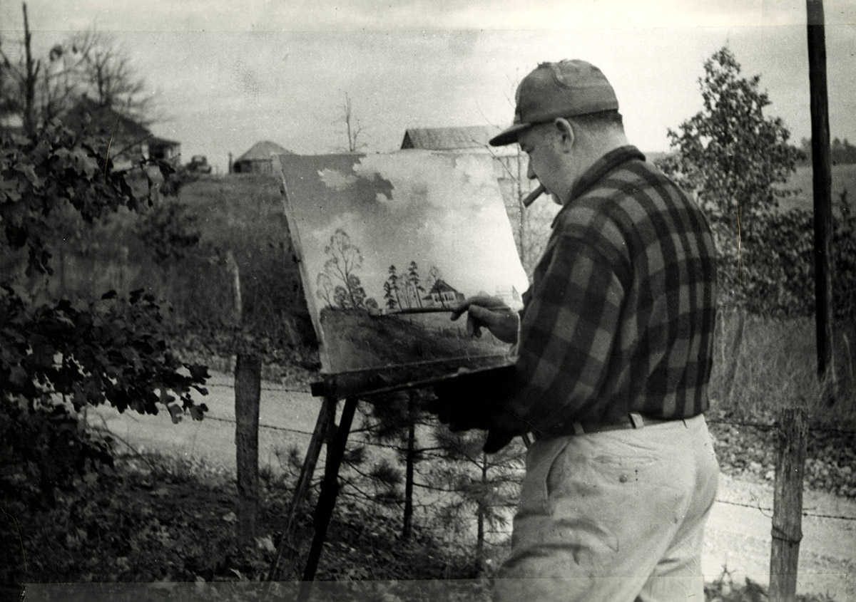 White man wearing a baseball cap with a cigar in his mouth painting at easel outdoors