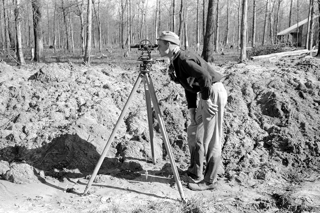 White man using surveying equipment in wooded area