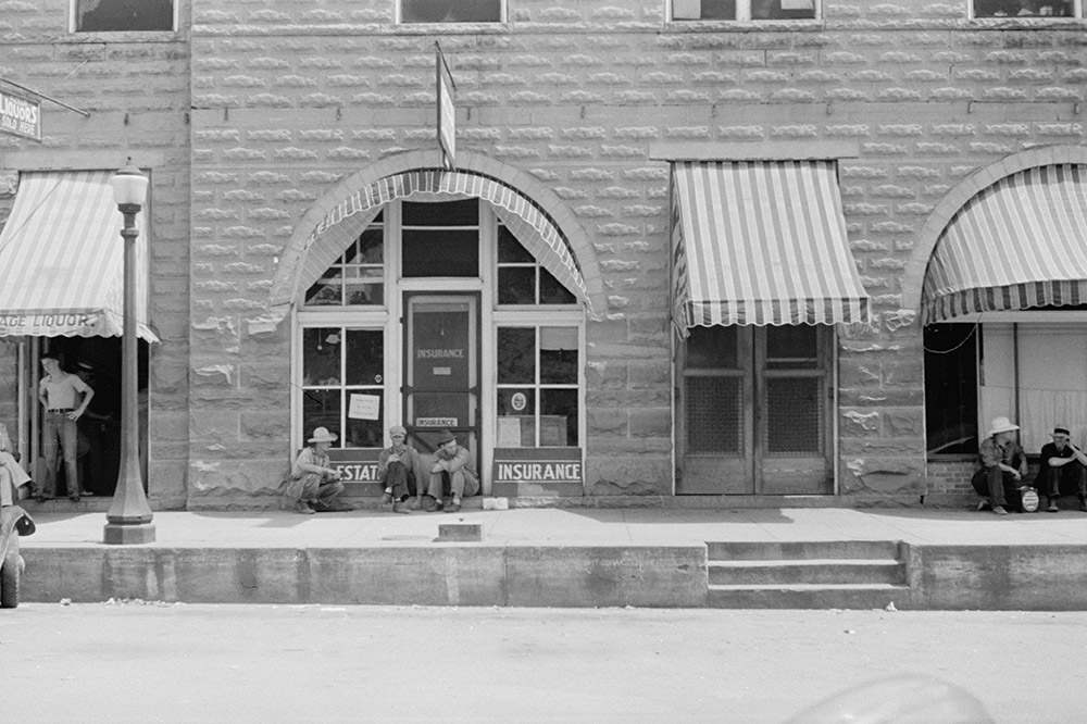 White men standing and sitting on sidewalk in front of multistory stone building
